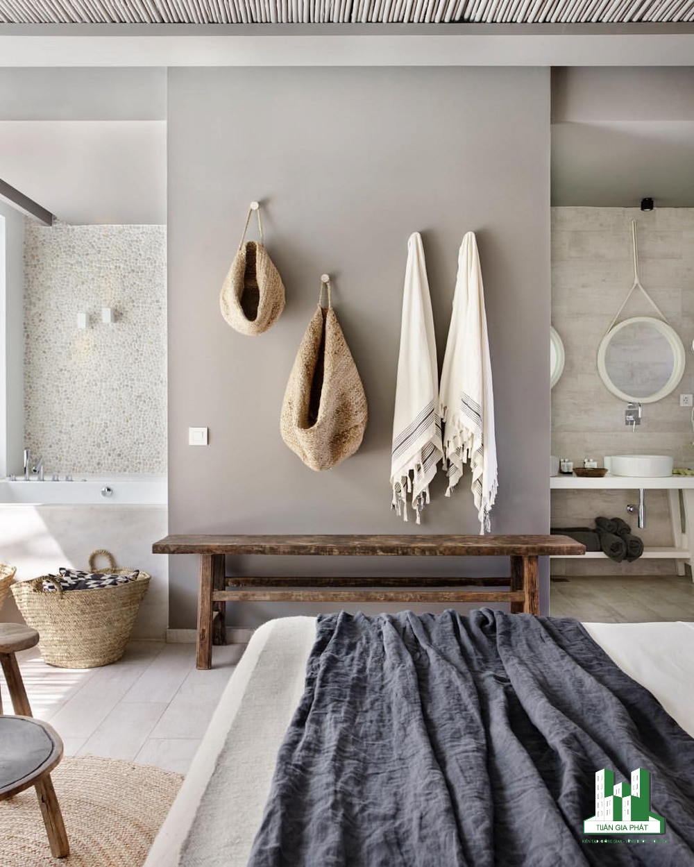 Phòng ngủ kết hợp với một phòng tắm bằng những bức tường đá và bê tông, một băng ghế gỗ, bộ đồ giường màu trung tính và những chiếc giỏ để lưu trữ.