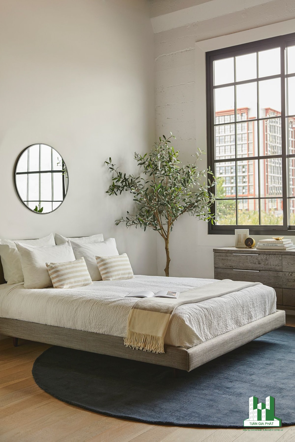 Phong cách hiện đại với giường nổi màu xám, tủ đựng quần áo bằng gỗ cũng tone màu này, cây thông, bộ khăn trải giường màu trung tính và chiếc gương tròn.
