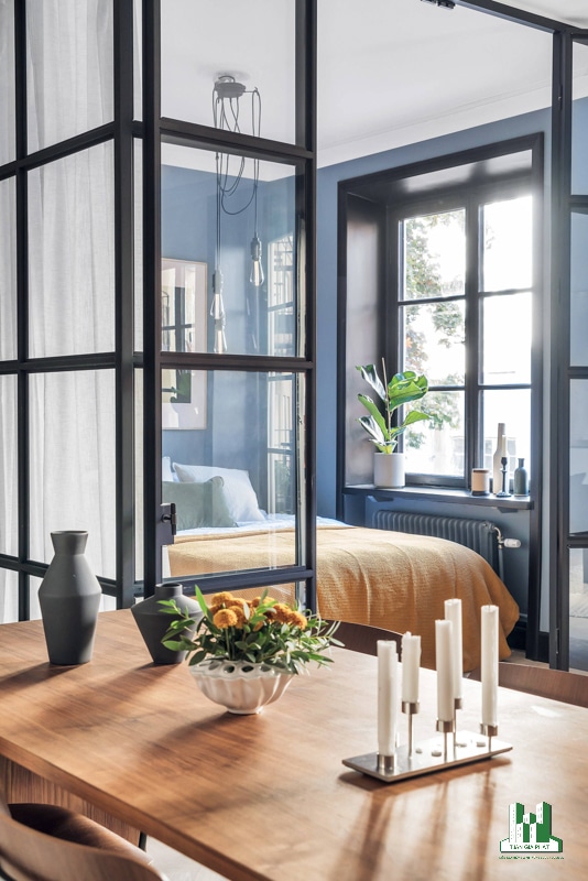 Phòng ngủ hiện đại với những bức tường xanh lam, một chiếc giường với vỏ chăn màu nắng, một số chậu cây, lọ hoa và những bức tường kính giúp tối đa hóa ánh sáng ban ngày.